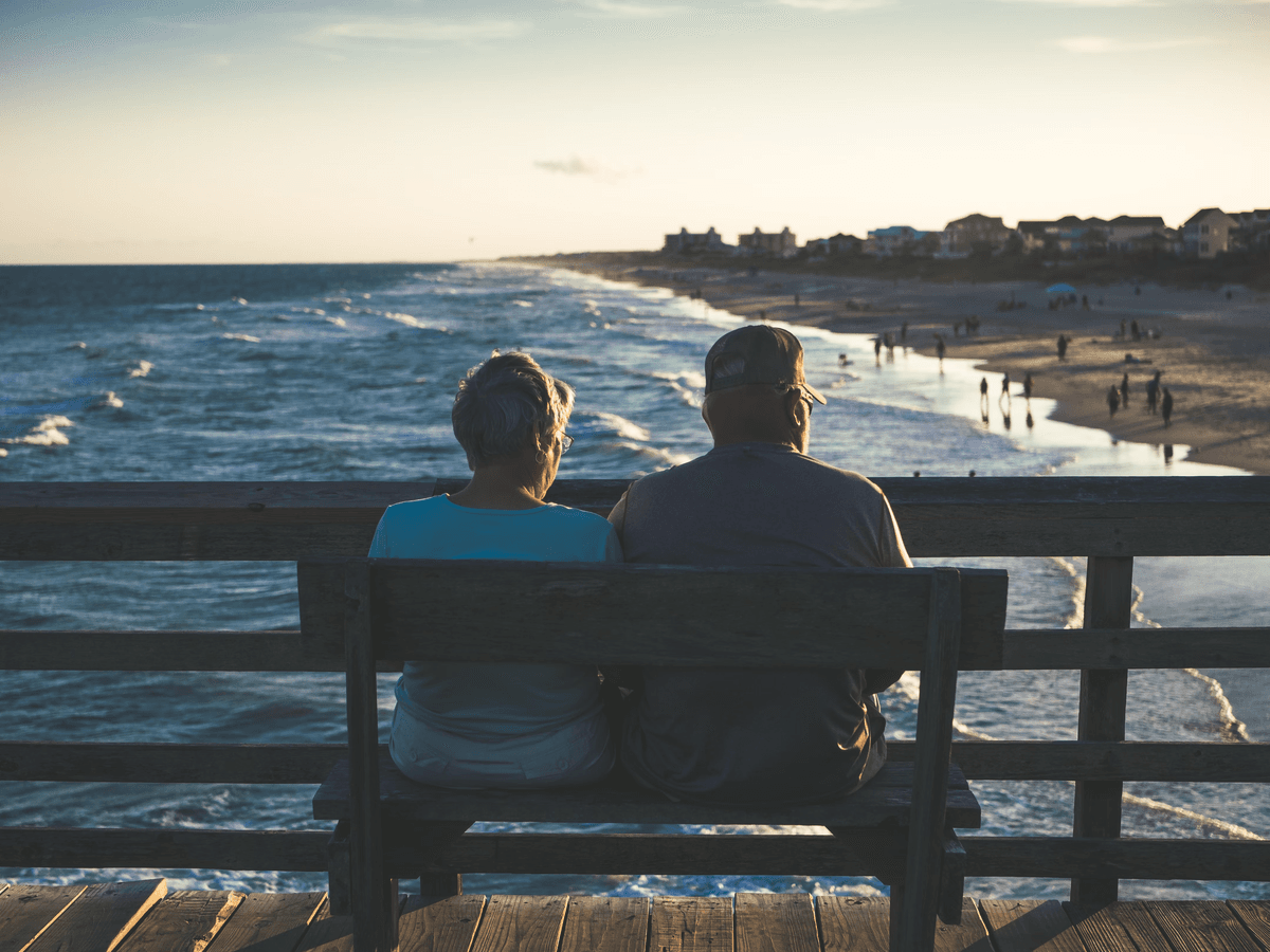 An elderly couple sits on a bench overlooking a beach at sunset, with waves gently lapping the shore and people walking in the distance.